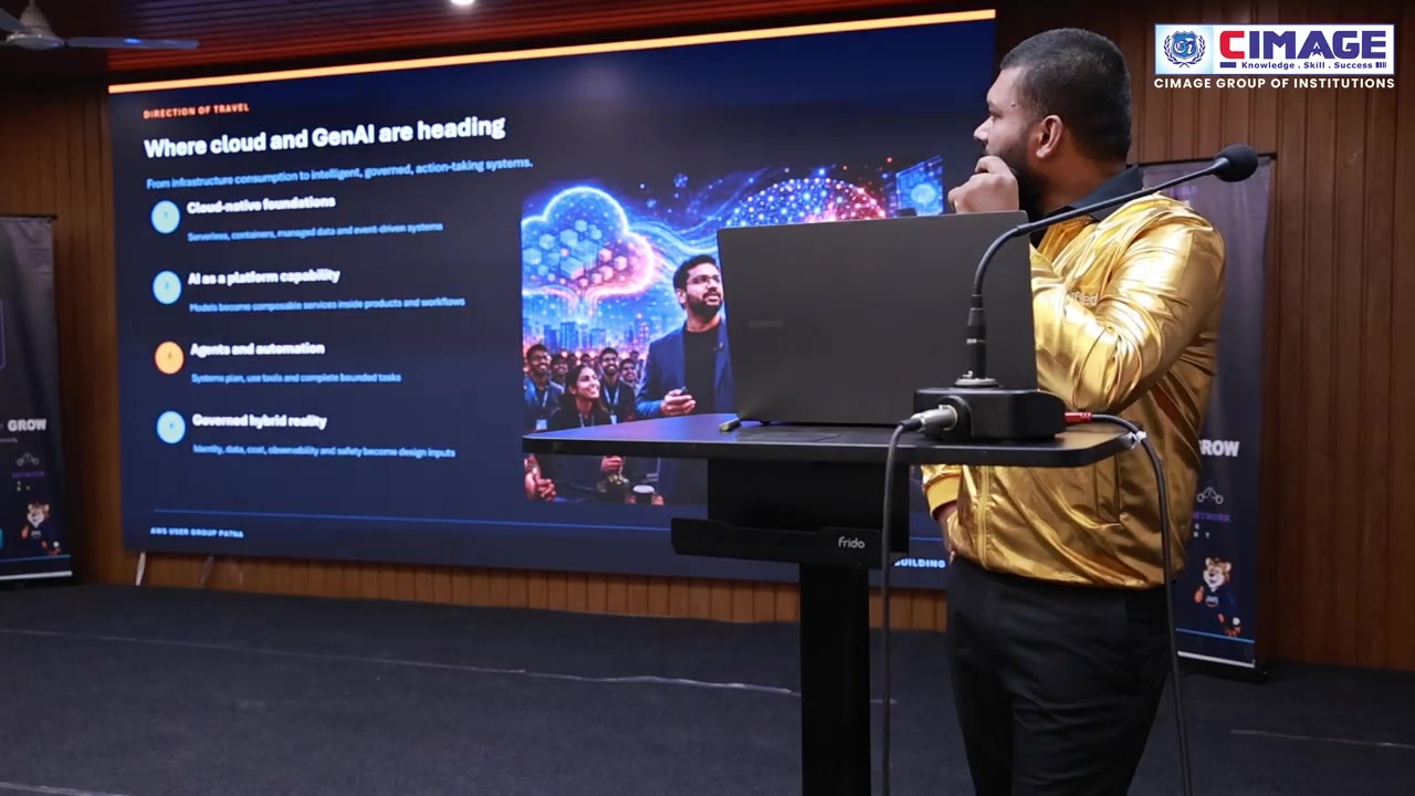 A speaker in a gold jacket stands on stage next to a podium facing a large presentation screen about cloud and GenAI.