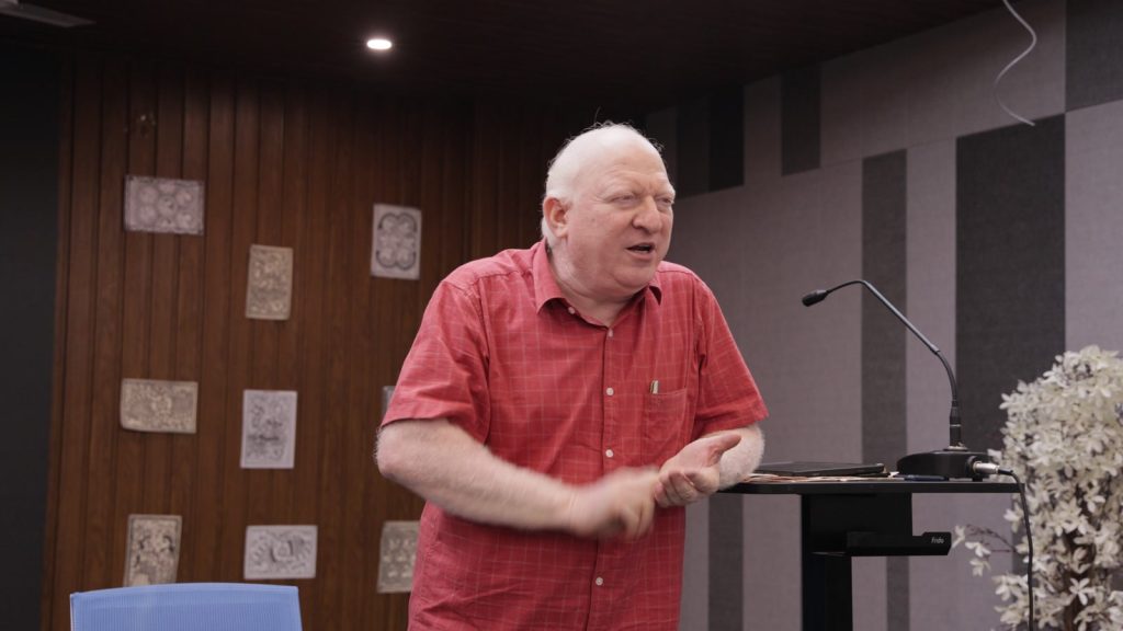A speaker in a red checkered shirt explains a concept with hand gestures in front of a wooden panel.